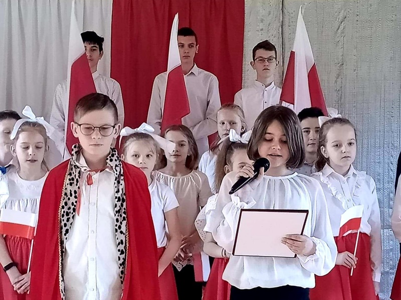 Uczniowie prezentują program artystyczny. Ubrani są w stroje biało-czerwone. Z tyłu trzech uczniów z polskimi flagami