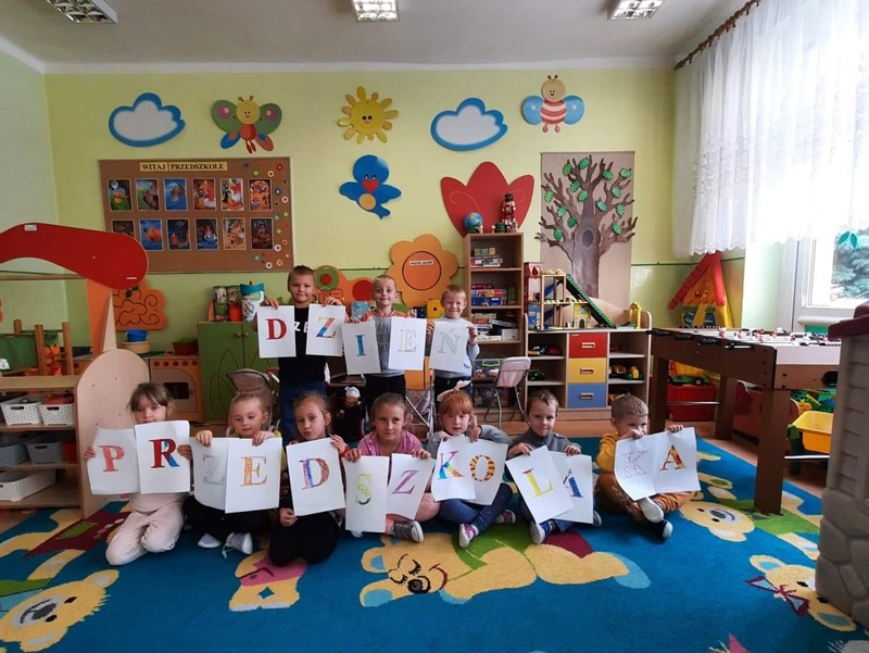 W pięknie udekorowanej  sali lekcyjnej dzieci siedzą na kolorowym dywanie. Trzymają przed sobą kartki z literami, które tworzą napis: Dzień Przedszkolaka
