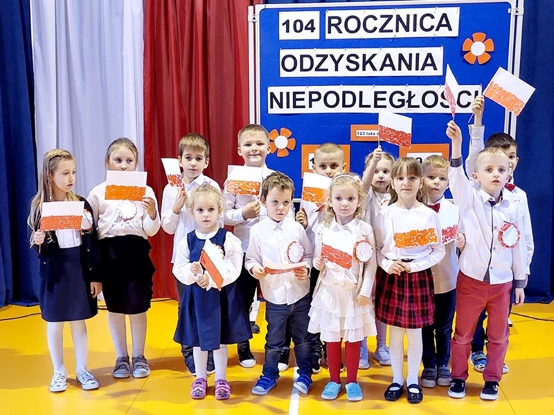Grupa najmłodszych uczniów z biało-czerwonymi chorągiewkami. Za nimi flaga narodowa i napis na niebieskim tle: 104 rocznica odzyskania niepodległości.