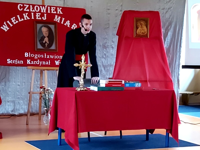 Uczeń grający Księdza  Kardynała stoi za czerwonym stołem, na którym znajduje się krzyż. W tle napis: Człowiek Wielkiej Miary