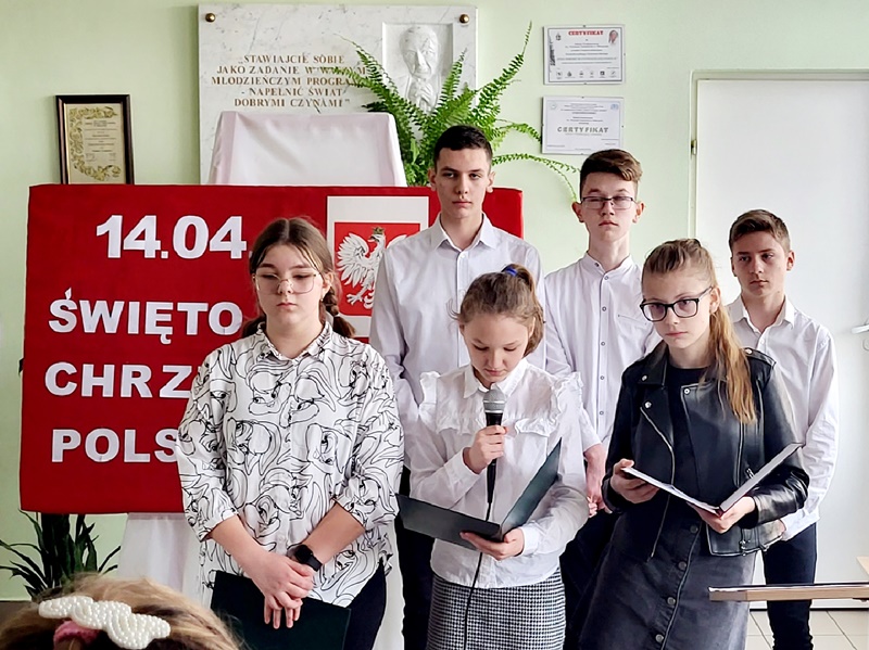 Na pierwszym planie grupa uczniów, którzy prezentują program artystyczny.  Za nimi napis informujący  o dacie chrztu Polski - biała czcionka na czerwonym tle.