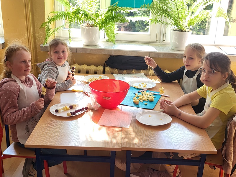 Czworo dzieci siedzi przy stoliku i przygotowuje owocowe szaszłyki 