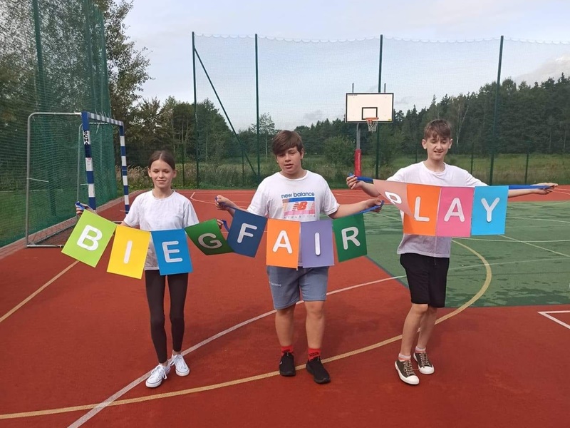 Trzech uczniów na boisku sportowym trzyma kolorowy napis: Bieg Fair Play