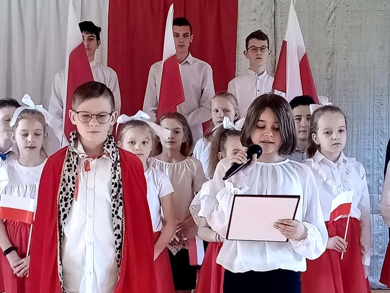 Uczniowie prezentują program artystyczny. Ubrani są w stroje biało-czerwone. Z tyłu trzech uczniów z polskimi flagami