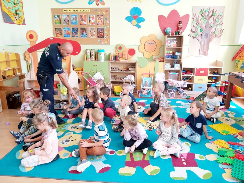 Policjant prowadzi zajęcia z dziećmi w oddziale przedszkolnym. Siedzą na dywanie w sali lekcynej.