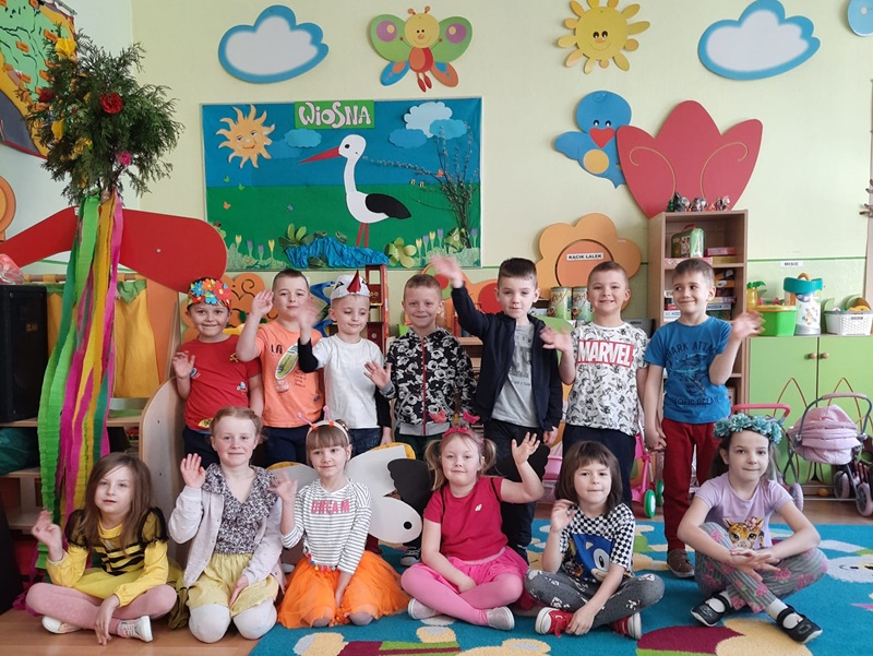 Dzieci ubrane w kolorowe stroje w sali lekcyjnej. Za nimi obrazek z bocianem na łące.